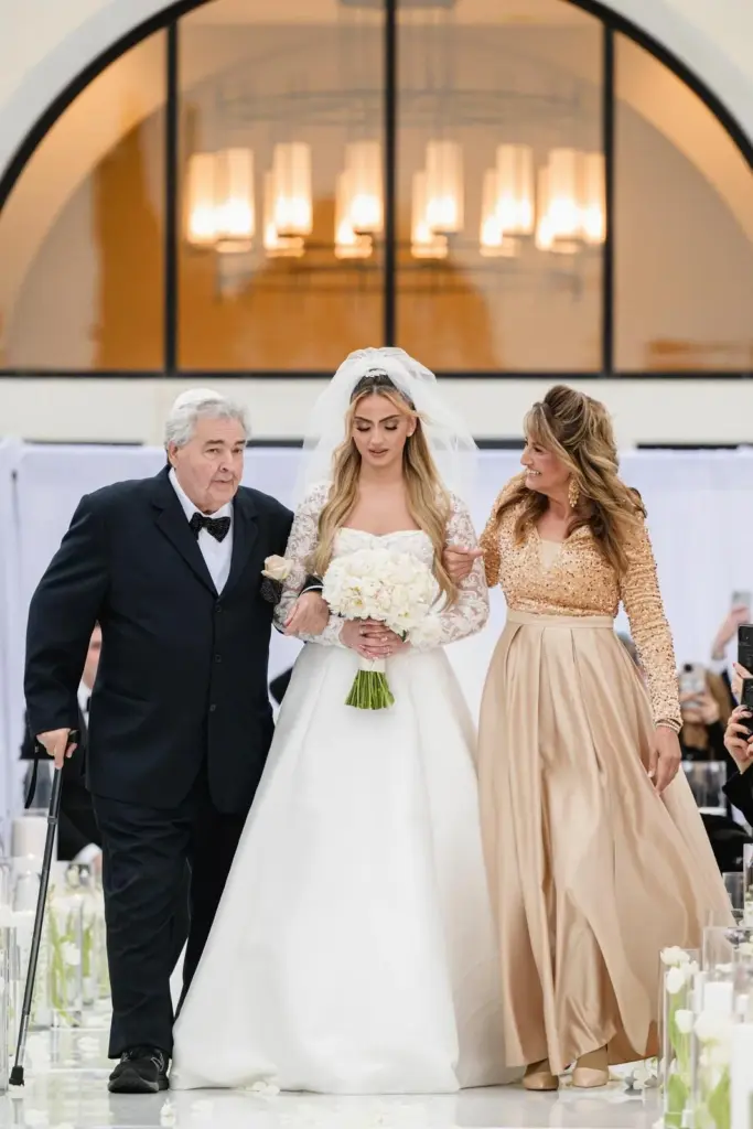 Bride walking down the aisle with her parents past an arched window and candlelit path