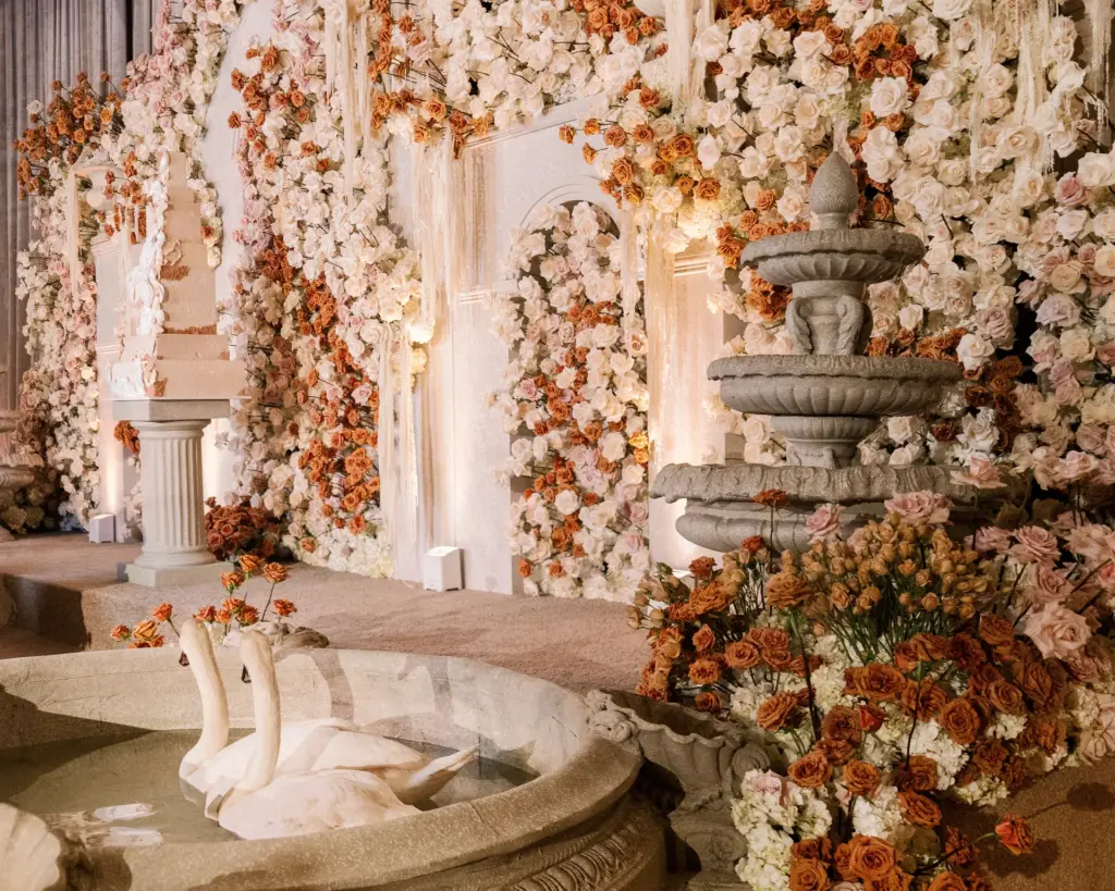 Floral wall backdrop with a wedding cake and a stone fountain featuring swan sculptures