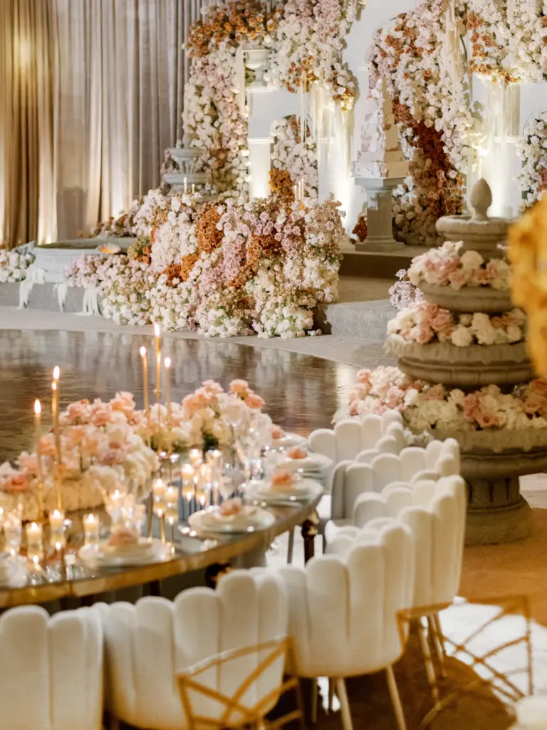 Wide view of a reception hall with a floral arch stage and candlelit scalloped chairs