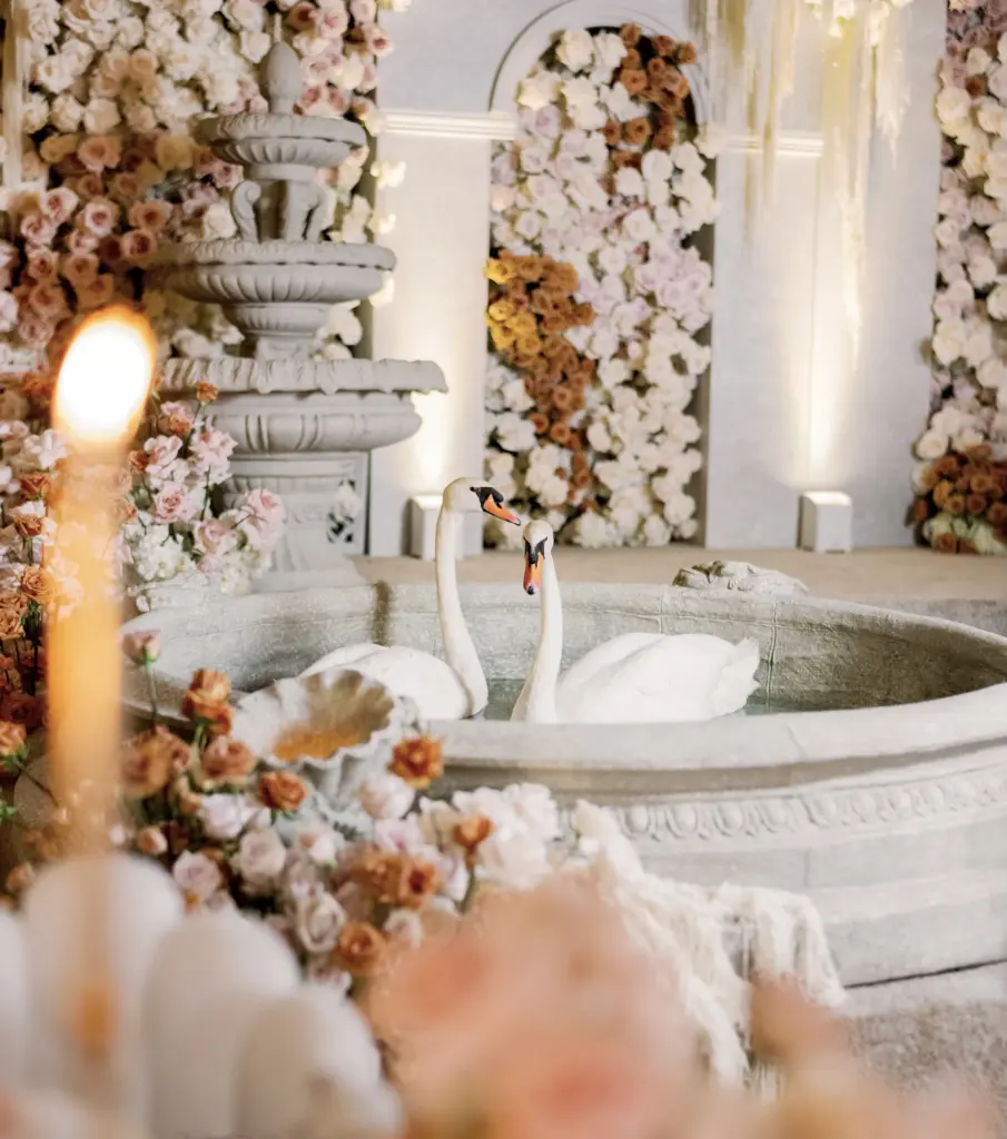 Close-up of a stone fountain with swan sculptures amid a floral wall backdrop