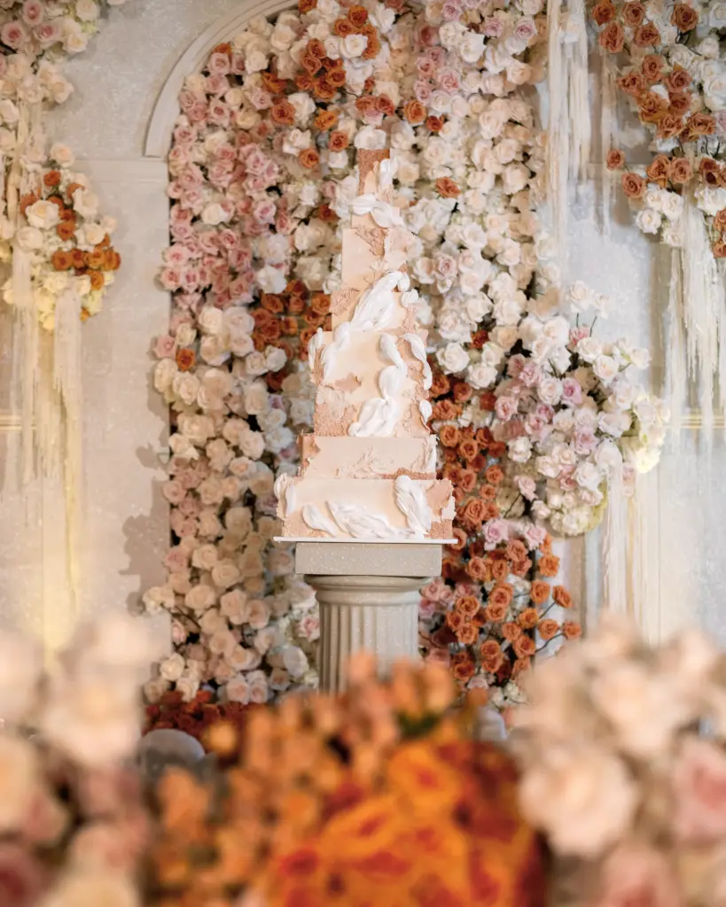 White wedding cake with sculptural draping displayed on a pedestal against a floral wall
