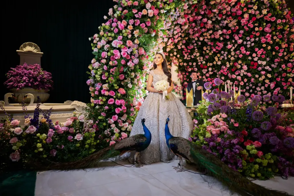 Bride in a beaded gown standing before a floral wall backdrop flanked by two live peacocks