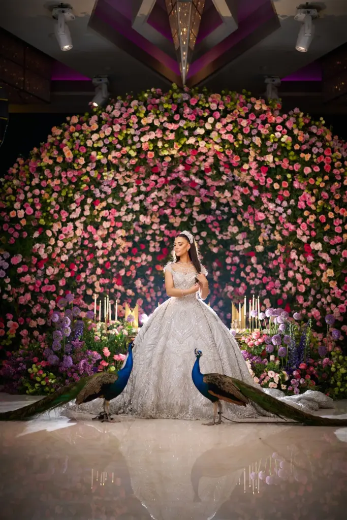 Bride in a sparkling ball gown posing before a rose flower wall with two peacocks