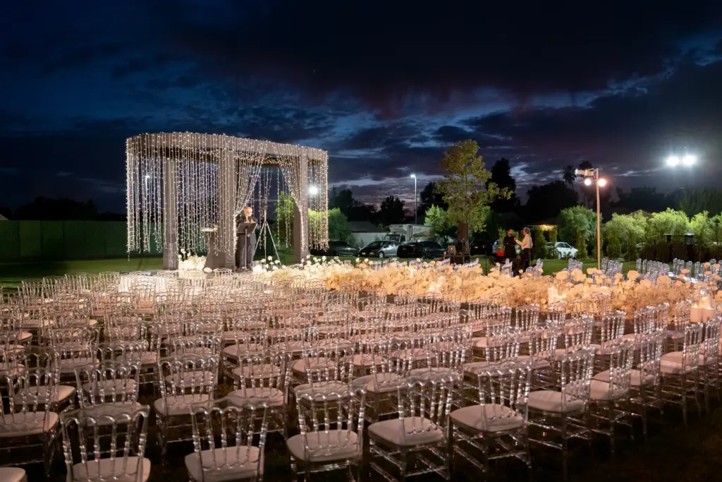 Outdoor evening wedding ceremony with a string-light gazebo and clear chiavari chairs