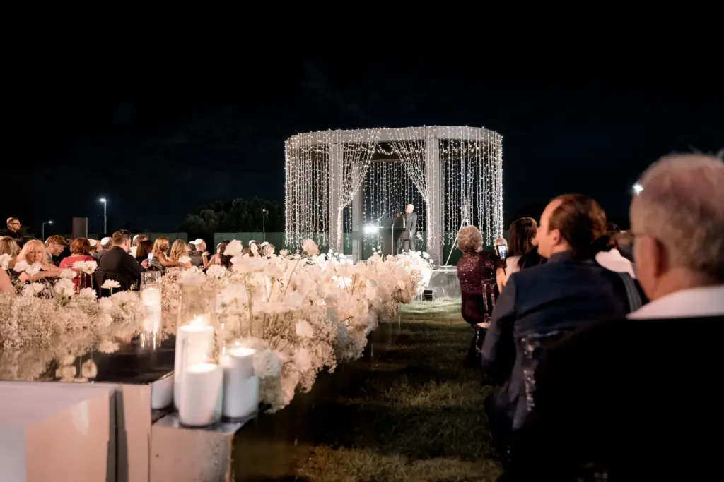 Guests seated along a white floral aisle at an outdoor evening wedding ceremony