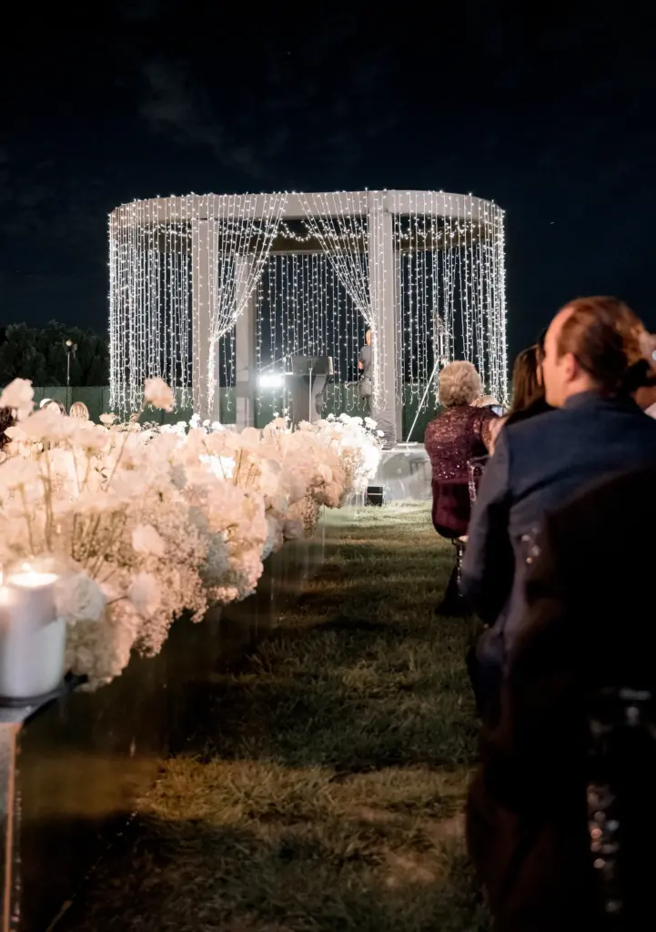 String-light gazebo at an outdoor wedding ceremony with a white floral aisle