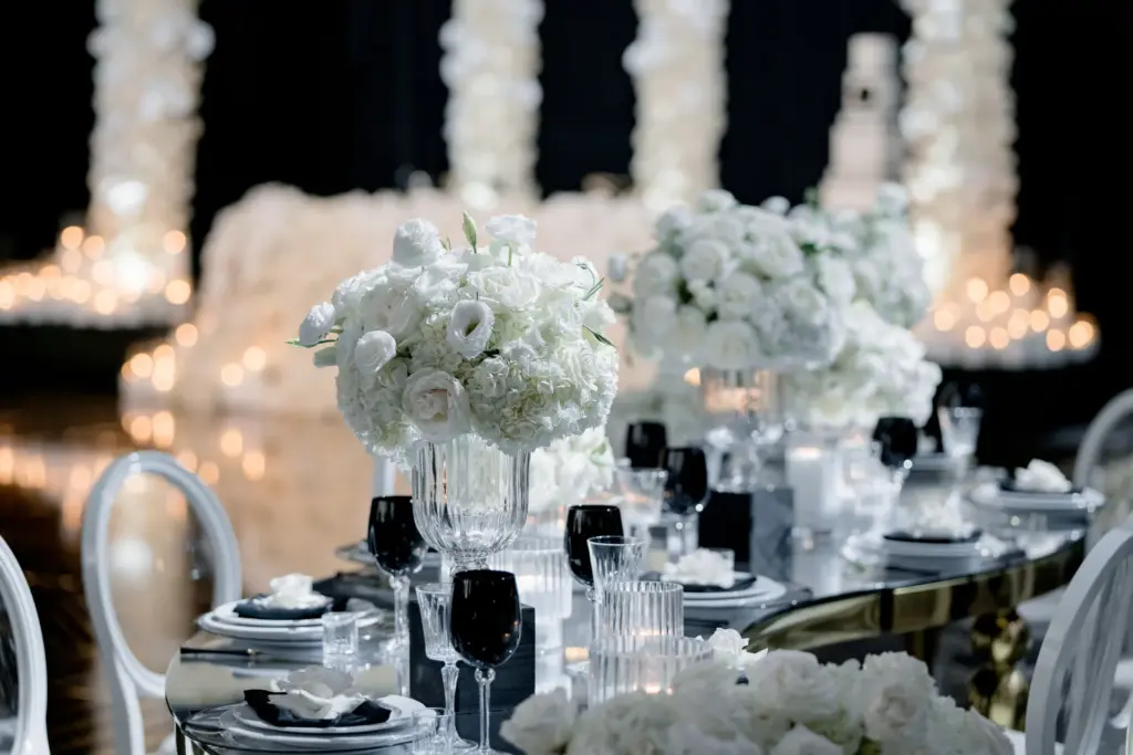 Table setting with a white floral centerpiece, black stemware, and tiered cakes in the background