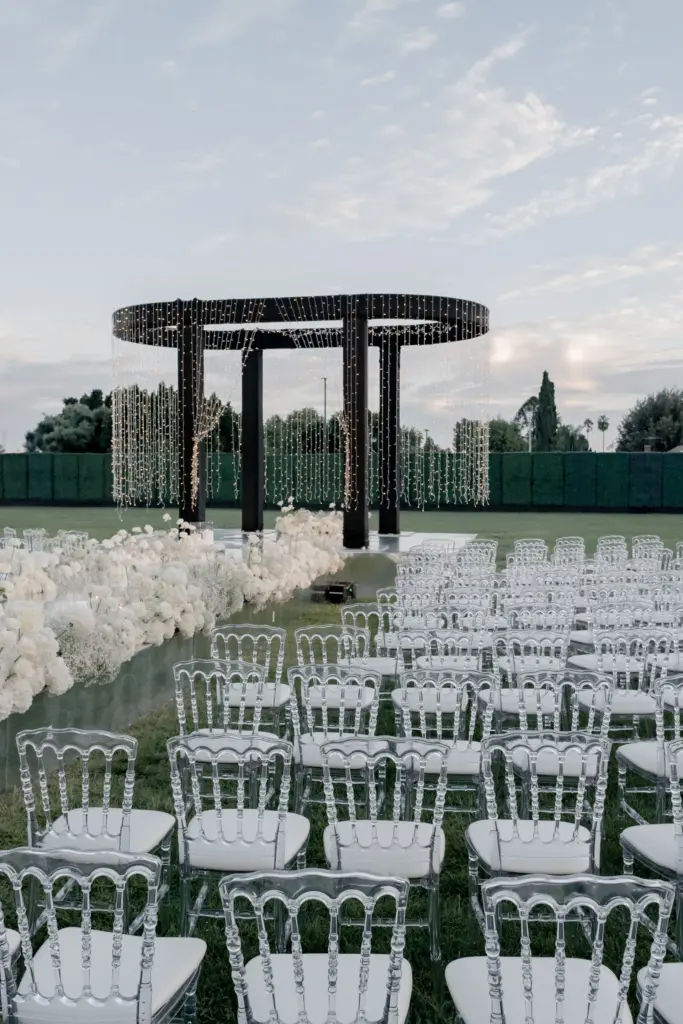 Outdoor wedding ceremony with a modern black ring arch and clear chiavari chairs at twilight